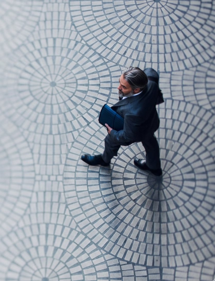 A grey-haired, bearded man in a suit walking across a city square, seen from above